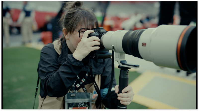 Photographer with telephoto lens mounted on tripod, capturing action at a sports field.
