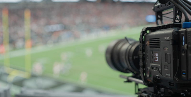 Sony cinema camera on a rig pointed at a blurred stadium field with crowds in background.