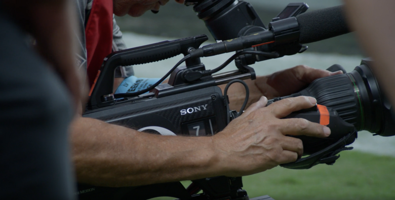 Cameraman operating a Sony professional video camera with a telephoto lens and microphone mounted on top.