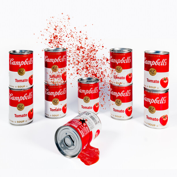 Campbell's soup cans arranged with one tipped over and red liquid splashing upward in a studio product photograph.