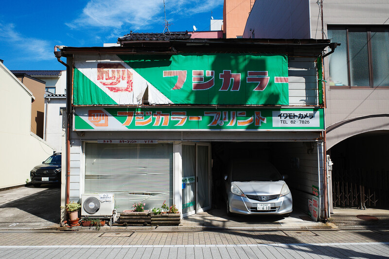 Abandoned Japanese car service garage with green signage and parked white vehicle under bright daylight.