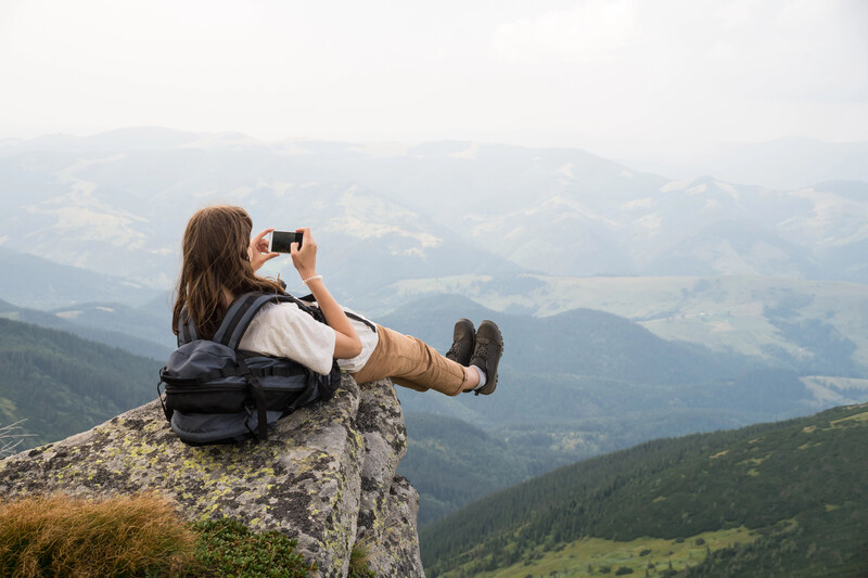 Hiker sitting on a mountain outcrop photographing the layered valley landscape below.