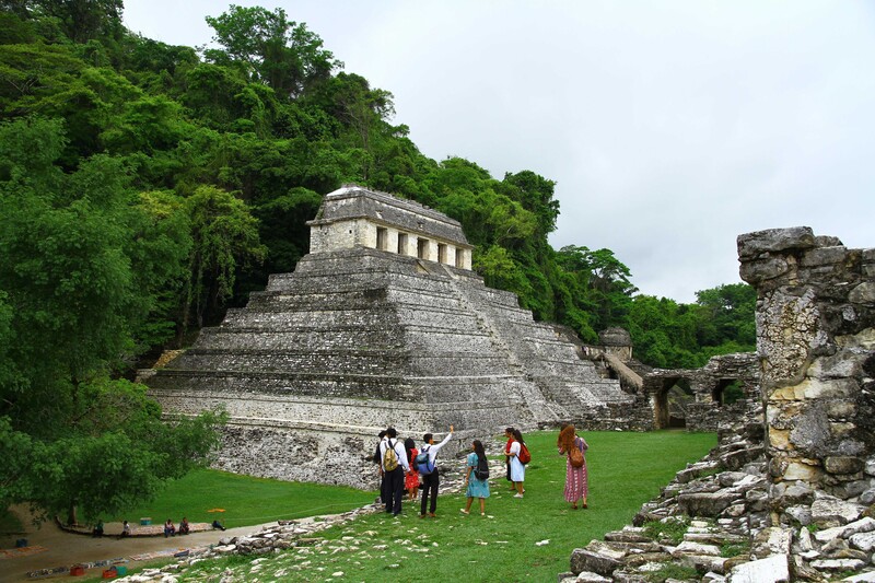 Ancient stone pyramid with temple structure atop, surrounded by lush green forest and visitors on grassy plaza.