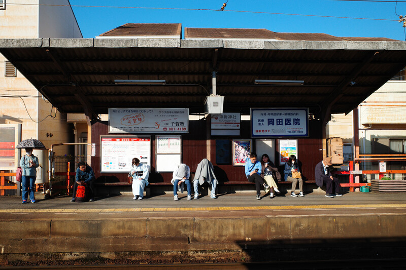 Japanese train station platform with passengers waiting under a covered shelter on a clear day.
