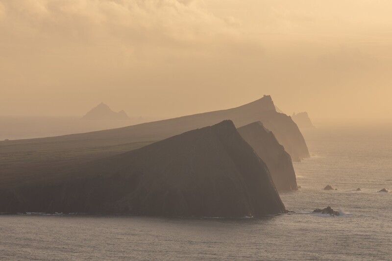 Dramatic coastal cliffs shrouded in thick fog at golden hour, with layered rock formations extending into misty ocean.