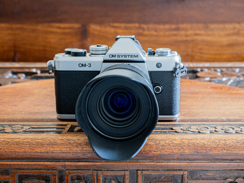 OM System OM-3 film camera with lens hood on wooden surface.