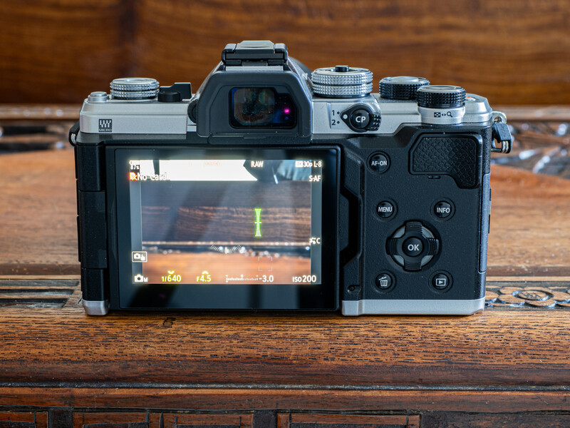 Mirrorless camera with interchangeable lenses displayed from rear, showing LCD screen with exposure histogram and control buttons on wooden surface.