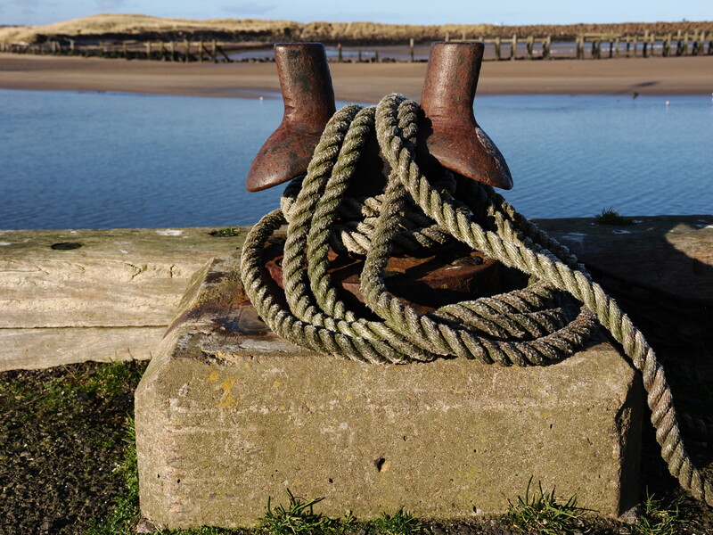 Weathered rope coiled around a rusty metal cleat on a concrete dock overlooking blue water.