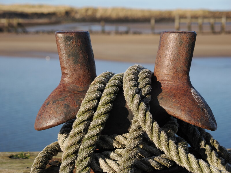 Weathered mooring bollards with coiled rope in foreground, calm tidal flat and distant pier beyond.