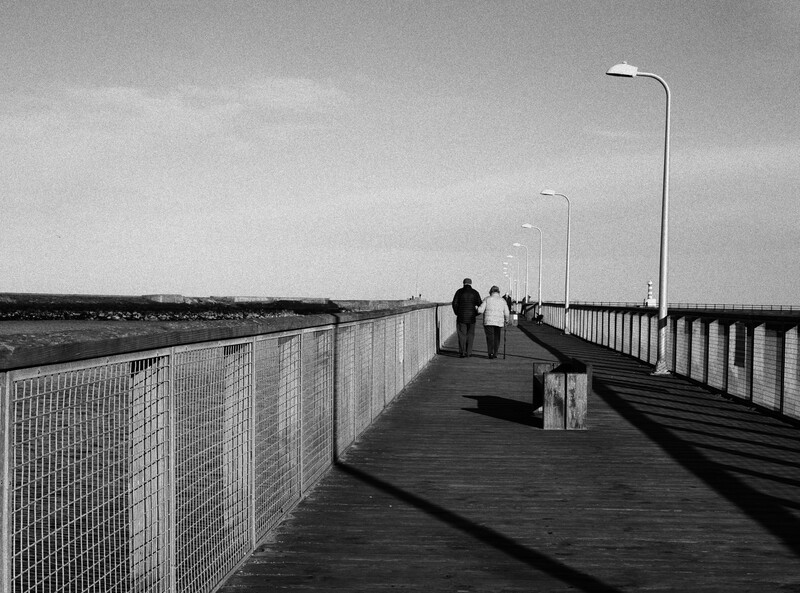 Two figures standing on a long wooden pier with metal railings extending toward the horizon under an overcast sky.
