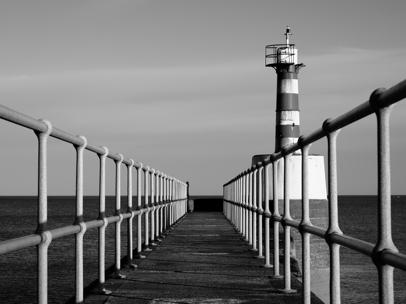 Wooden pier with white railings stretching toward a striped lighthouse under overcast sky.