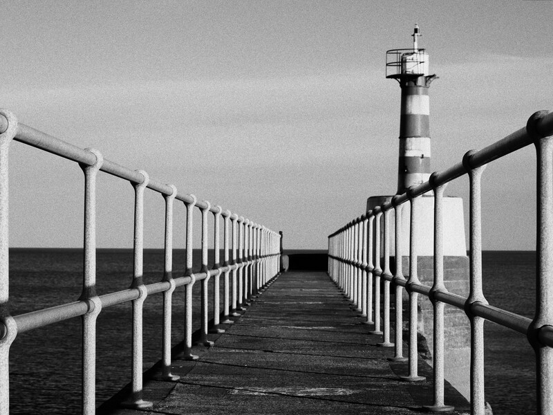 Symmetrical black and white photograph of a pier with metal railings extending toward a lighthouse in the distance.
