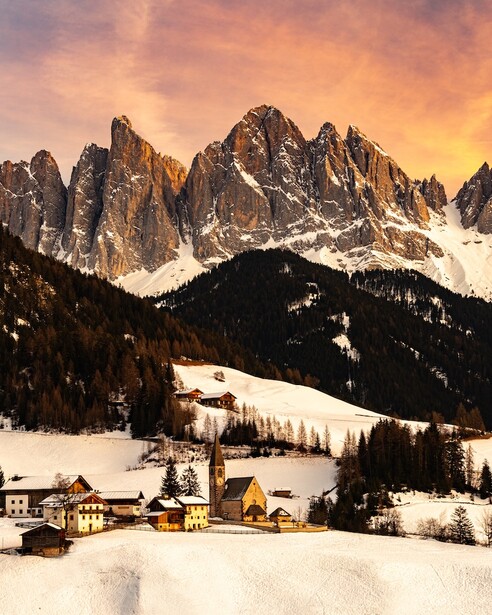 Snow-covered alpine valley with dramatic jagged peaks at sunset, small village nestled in forefront.