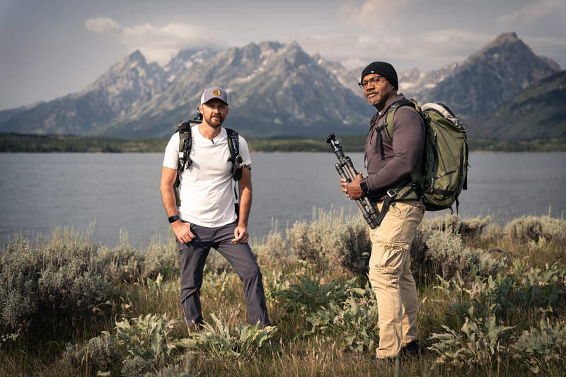 Two hikers with backpacks and camera gear stand in sagebrush landscape facing snow-capped mountains across a lake.