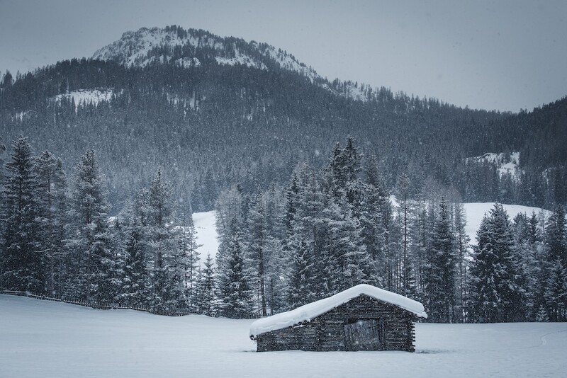 Stone cabin in a snowy meadow surrounded by forested mountains under overcast skies.