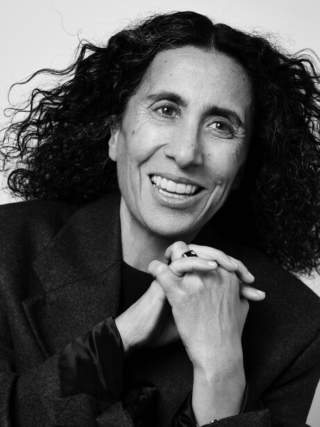 Black and white portrait of a person with long dark curly hair, smiling at the camera with hands clasped near chest.