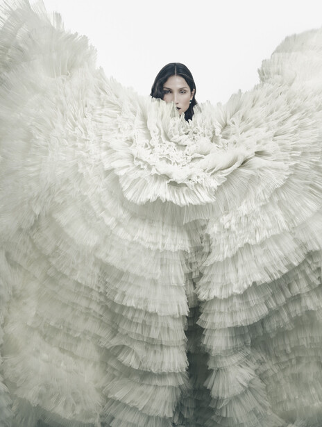 Person with dark hair wearing an elaborate white feathered wing costume against a pale, misty background.