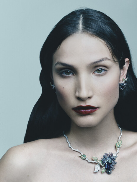 Portrait of woman with long dark hair and bold burgundy lipstick wearing ornate silver necklace against neutral background.
