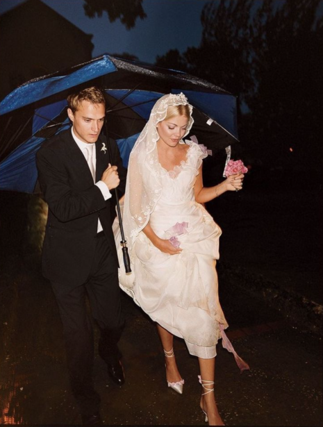 Couple sheltering under blue umbrella during nighttime rain, groom in dark suit and bride in white dress.