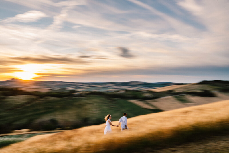 Two people standing on a hillside during golden hour sunset with rolling landscape below.