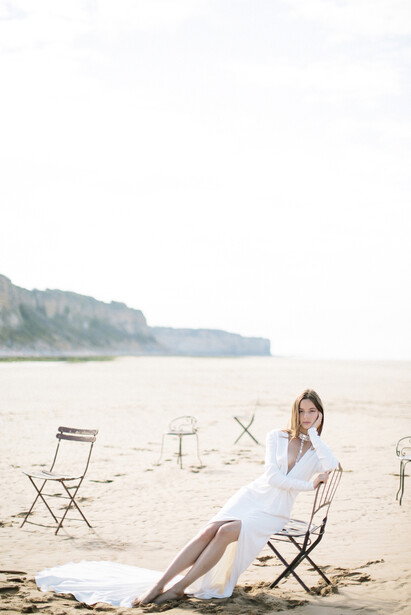 Woman in white dress seated on folding chair on sandy beach with calm water and distant coastline behind her.