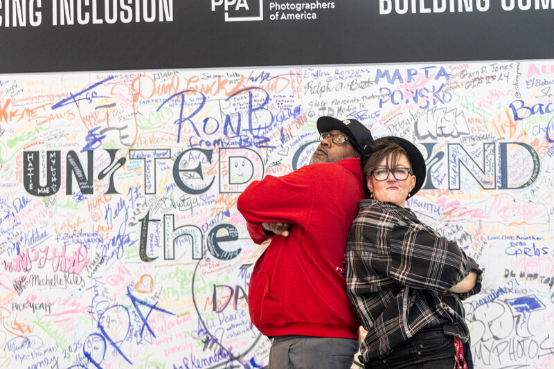 Two people posing back-to-back in front of a colorful wall covered with handwritten signatures and messages.