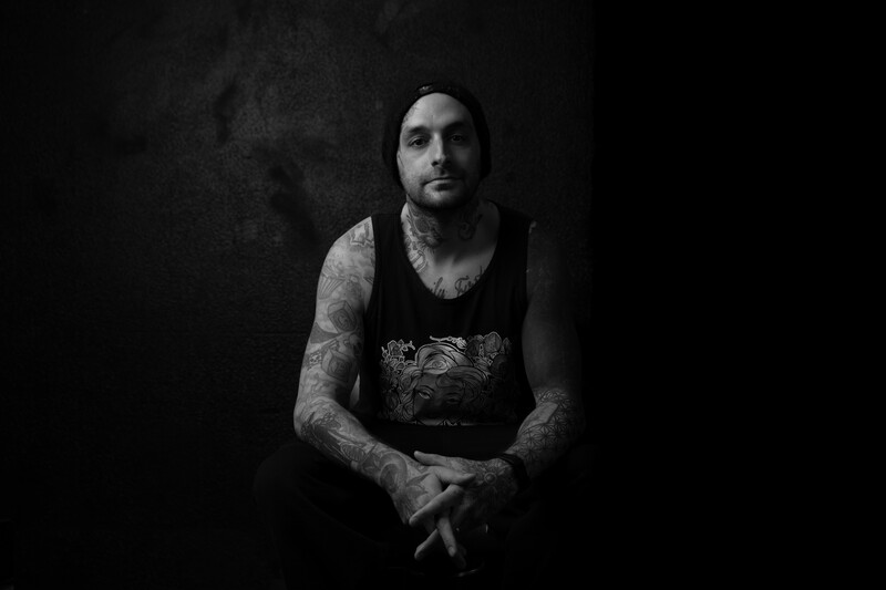 Tattooed man with dark hair wearing a black sleeveless shirt against a dark textured background.