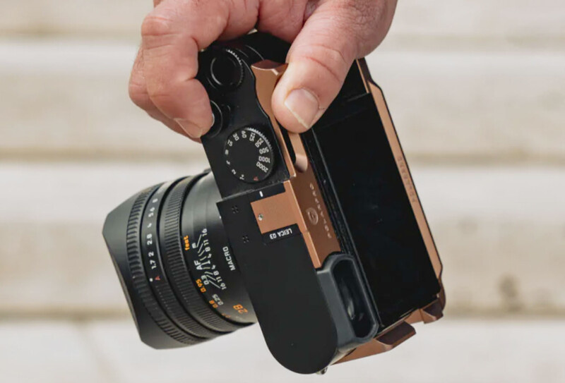 Hand holding a rangefinder camera with attached lens against a blurred brick wall background.