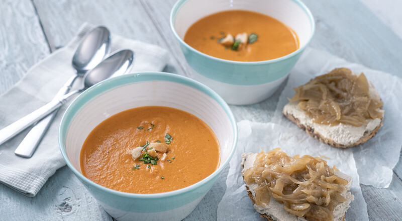 Two bowls of creamy orange soup garnished with herbs, served with sliced bread spread with white cream on a light blue wooden surface.