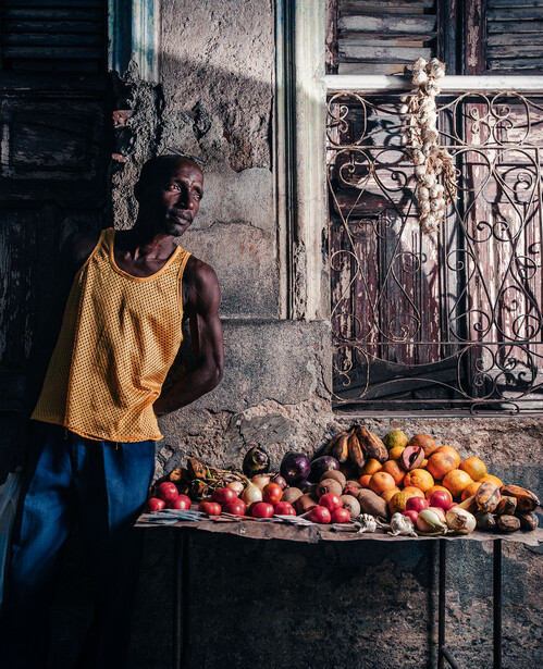 Man in yellow tank top standing beside a table displaying fresh tomatoes and other produce against a weathered building facade.