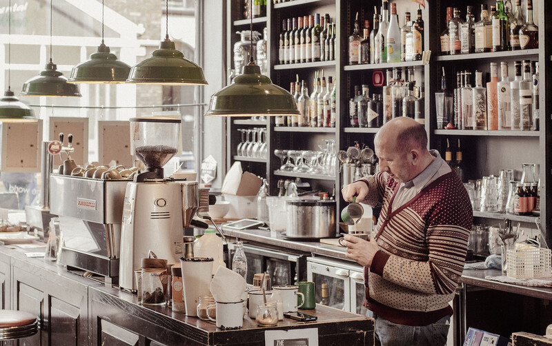 Barista working behind espresso machine in minimalist café with industrial pendant lights and shelved product display.