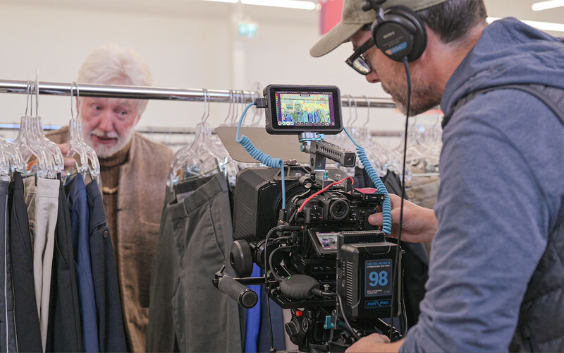 Cinematographer operating a professional cinema camera while interviewing an older man next to clothing racks.