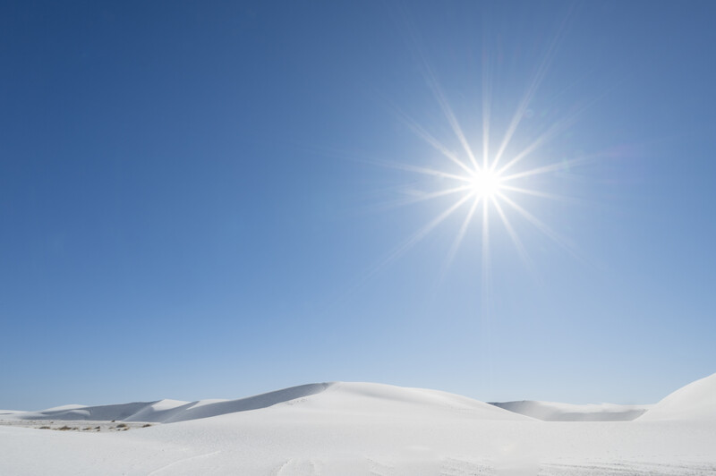 Bright sun casting lens flare over white sand dunes under clear blue sky.