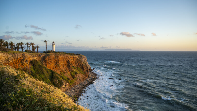 Lighthouse on a clifftop overlooking the ocean at sunset, with palm trees and golden-hour lighting.