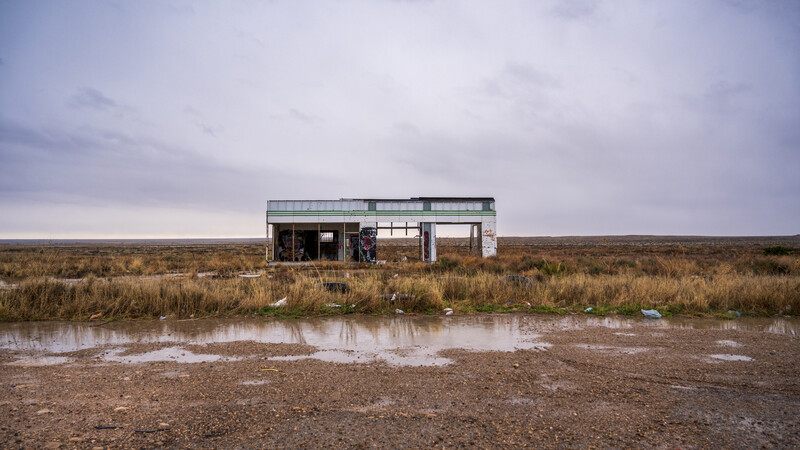 Abandoned single-story corrugated metal structure standing alone in flat, barren marshland with shallow water in foreground.