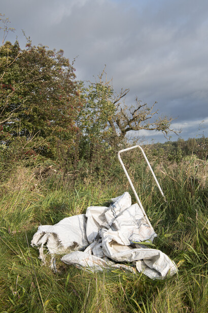 Abandoned white baby stroller in overgrown grass field beneath cloudy sky.