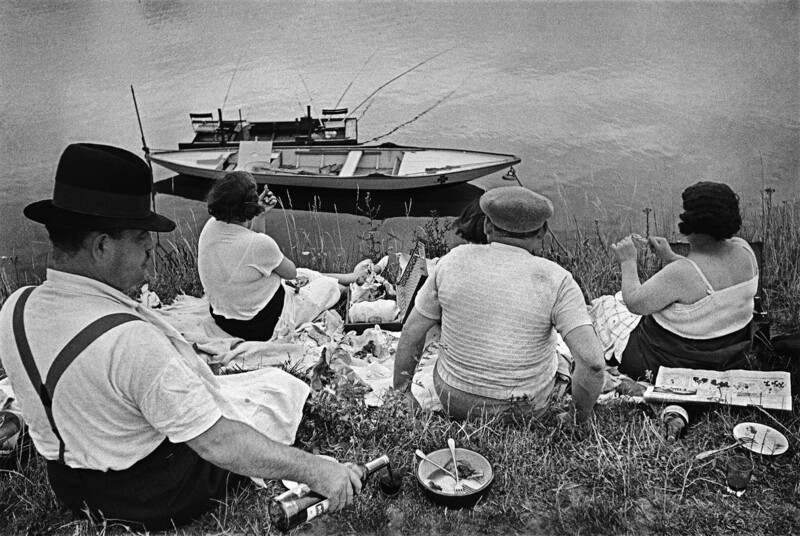 Four people having a picnic on grass beside a moored boat in black and white.