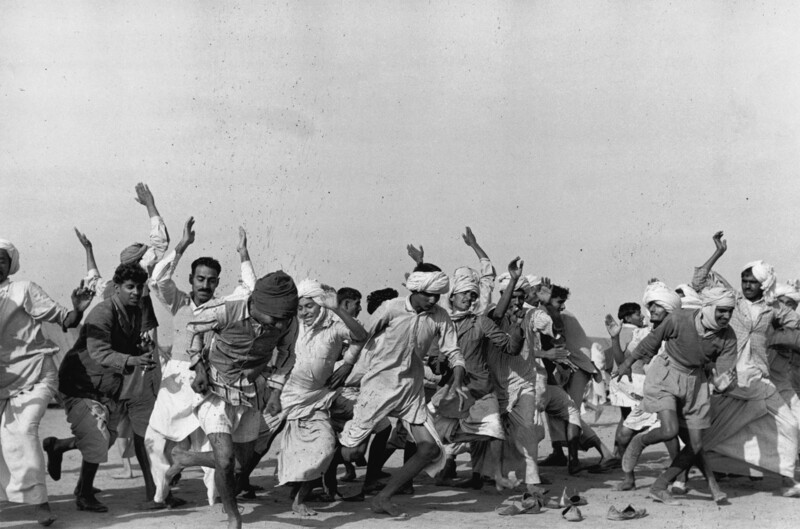 Group of people in traditional clothing dancing with raised arms in dusty outdoor setting.