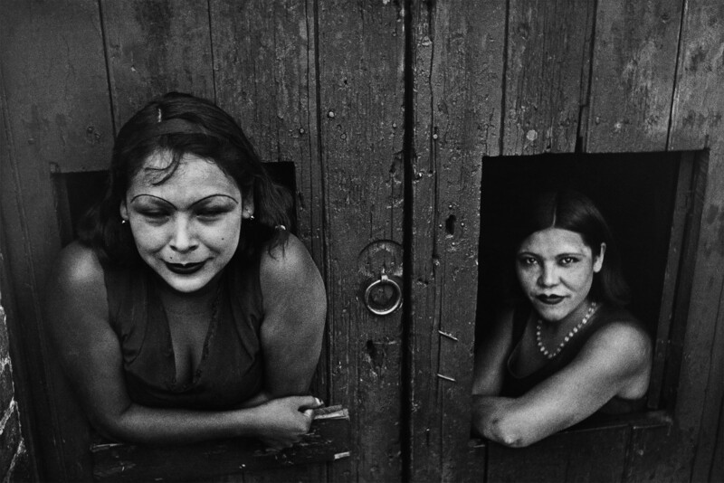 Two women peer through wooden doorways in a black and white portrait study.