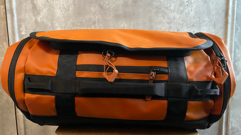 Orange and black cylindrical duffel bag with compression straps displayed against a concrete wall.