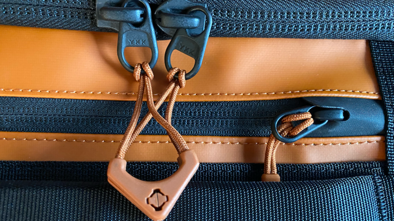 Close-up of a tan leather camera bag strap with blue dual carabiners and a heart-shaped security lock.