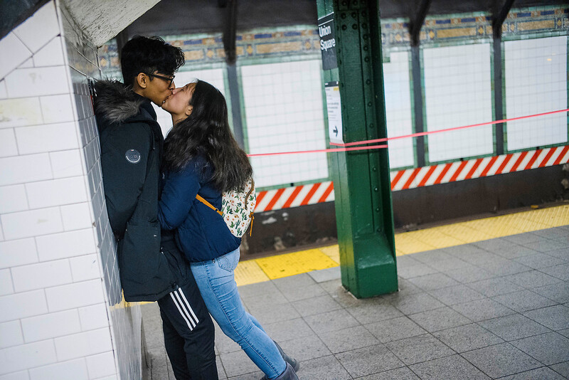 Couple kissing against a white tile wall at a subway station platform.