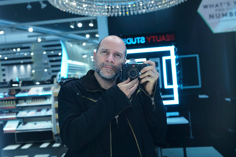 Photographer holding a camera in a modern retail electronics store with neon signage and bright overhead lighting.
