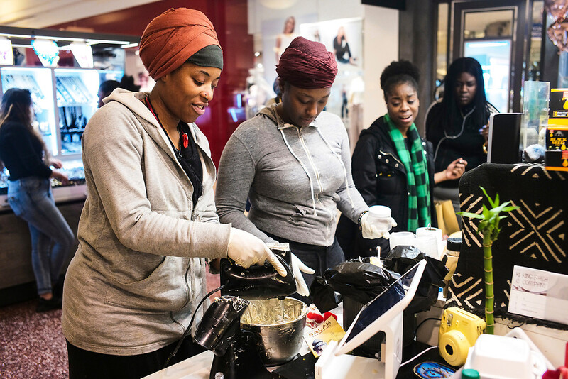 Two women in head wraps work together at a community event station with cooking equipment and supplies.