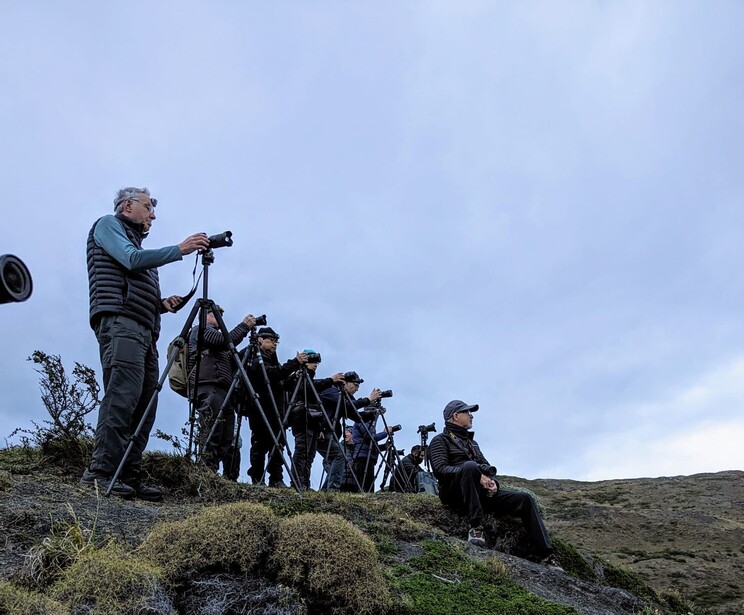 Photographer using a telephoto lens on a tripod while standing on rocky terrain with other photographers lined up behind.