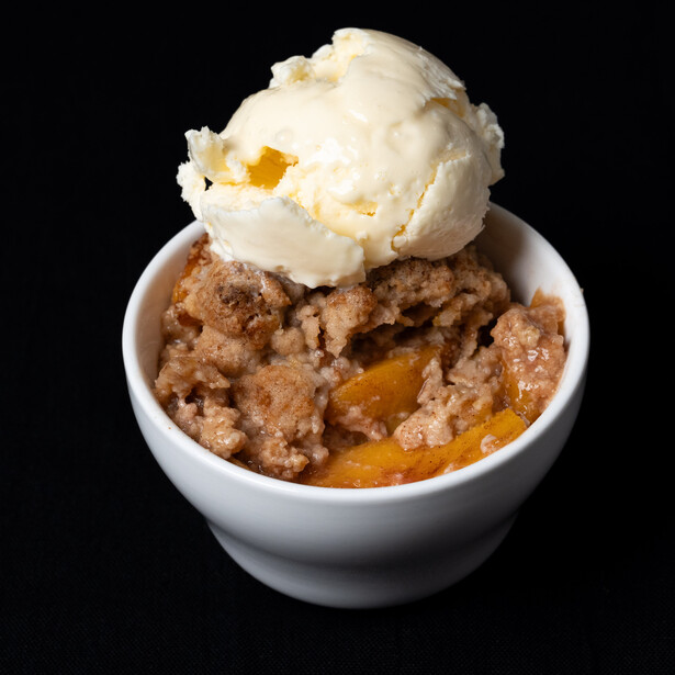Warm apple crisp topped with vanilla ice cream in a white ceramic bowl against black background.