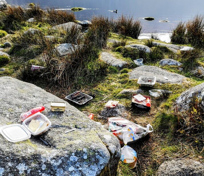 Scattered plastic waste and debris on rocky coastal terrain with sparse vegetation and water visible in background.