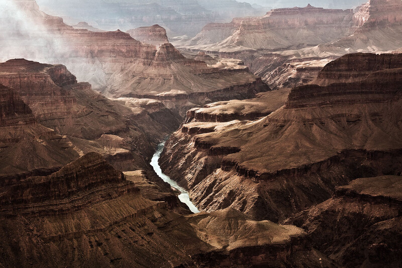 A winding river cutting through layered canyon walls under hazy sunlight. The Colorado River meanders through the Grand Canyon.