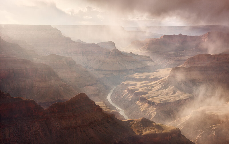 Grand Canyon landscape with layered rock formations, a winding river, and atmospheric haze illuminated by dramatic sunlight.