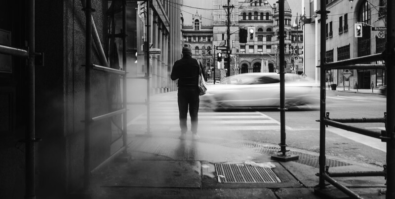 Man standing in urban alley with blurred vehicle passing on street beyond.